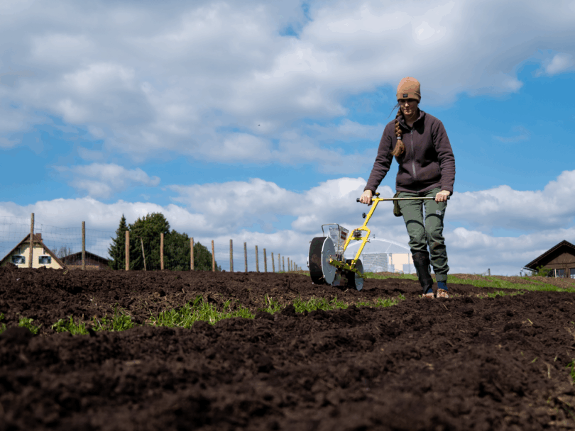 Nina mit einer handbetriebenen Sämaschine auf einer landwirtschaftlichen Fläche.