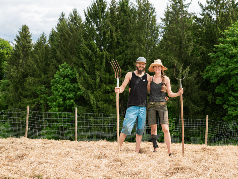Getrocknetes Heu liegt am Boden, Selim (links) und Nina (rechts) von der Wilden Ernte stehen Arm in Arm mit jeweils einer Heugabel und lachen in die Kamera.